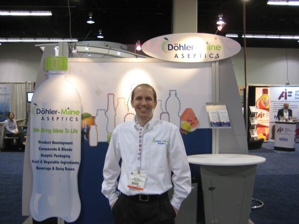 A smiling man in a white shirt and lanyard stands in front of the Döhler-Milne Aseptics booth, which has a large bottle-shaped sign listing product development and aseptic packaging services.