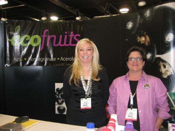 Two women wearing badges stand behind a table at the ecofruits booth. The black backdrop reads ecofruits, with açaí, pomegranate and acerola, and shows dark berries.