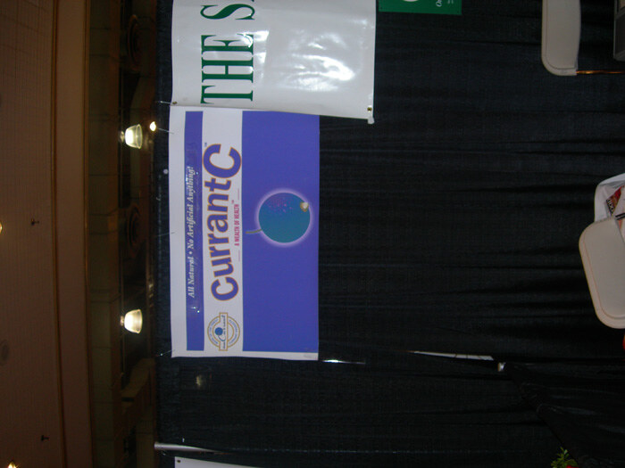 A sideways photo of a purple and white Currant C banner with a blue globe graphic, hung on a dark curtain. Other signage and folding chairs are partly visible.