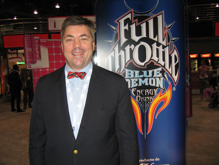 A smiling man in a dark blazer and red bow tie stands in front of a tall Full Throttle Blue Demon energy drink display in a convention hall.