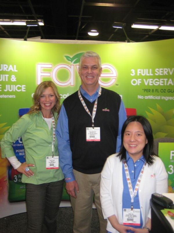 Two women and a man wearing badges and lanyards pose in front of a green Fave banner that mentions fruit and vegetable juice and three servings of vegetables.