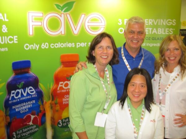 Four people, three women and a man, crowd together for a photo in front of a green Fave backdrop with bottles of Fave juice, including Pomegranate Blueberry Goji.