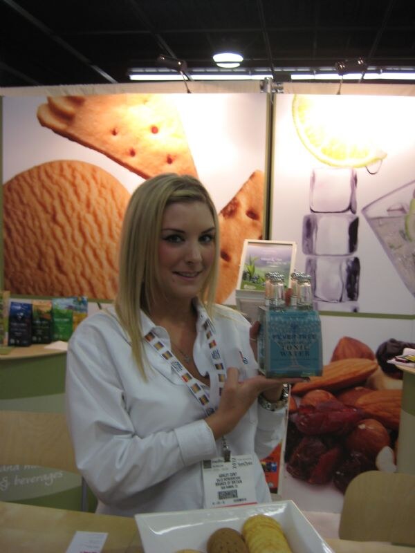 A blonde woman with a lanyard holds a four-pack of Fever-Tree tonic water in front of displays of biscuits, almonds and dried fruit, with a plate of cookies in the foreground.