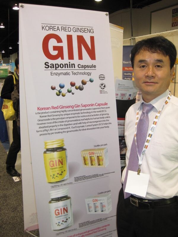 A man in a white shirt and purple tie stands beside a tall banner for GIN Saponin Capsule Korean red ginseng, which shows product bottles and boxes with descriptive text.