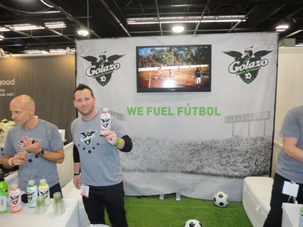 A man in a gray Golazo shirt holds up a bottle in front of a banner reading We Fuel Fútbol, with a TV above, artificial turf and soccer balls. Another man gestures beside him.