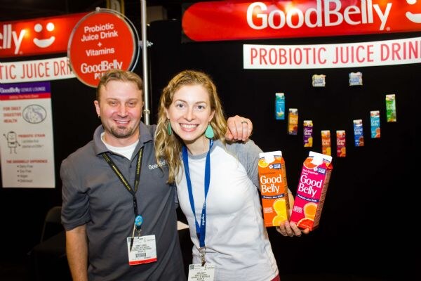 A smiling man in a gray polo and a smiling woman with long wavy hair hold cartons of GoodBelly probiotic juice drink in a booth with GoodBelly signs behind them.