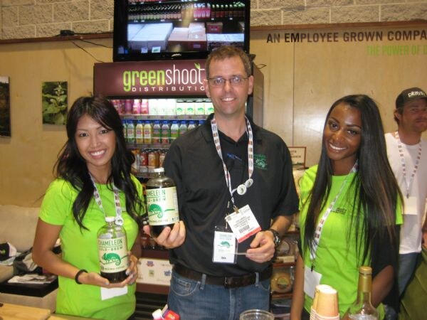 Three people stand at a Greenshoots Distribution display; a man in a black shirt and a woman in green hold bottles of Chameleon Cold-Brew, and a man stands behind them.