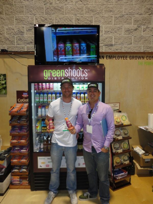 Two men in caps stand either side of a Greenshoots Distribution refrigerator, each holding a bottled drink. A CoGo snack rack and a screen showing beverages are nearby.