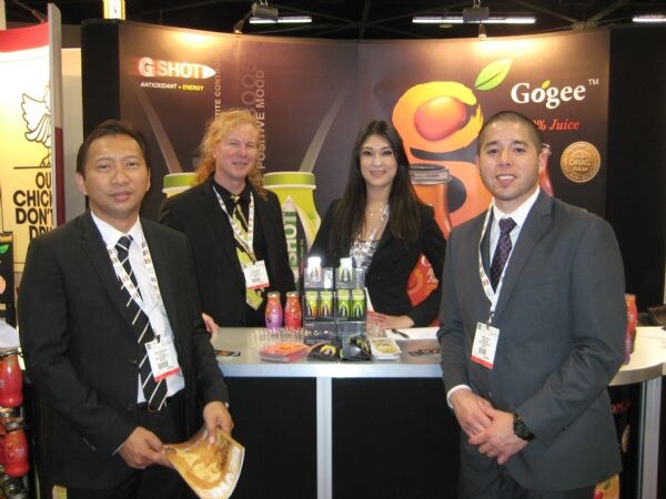 Two men in suits stand in front, with a man with long hair and a woman standing behind a counter, at a booth with G Shot and Gogee signs and bottles on display.