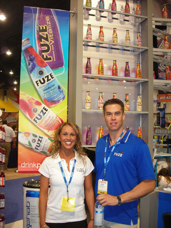 A smiling woman in a white top and a man in a blue Fuze polo, both wearing lanyards, stand before a Fuze banner and shelves of colorful bottled drinks.