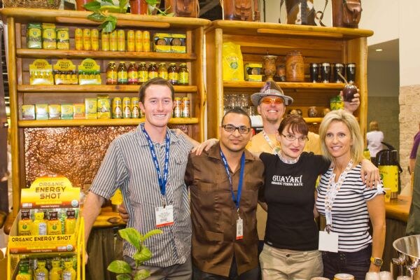 Four people stand close together in front of wooden shelves of bottled drinks at the Guayaki booth; an Organic Energy Shot display sits at left and a fifth person in a hat stands behind.
