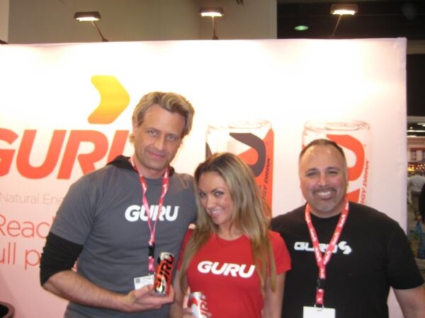 Two men and a woman wearing Guru shirts and lanyards pose in front of a white Guru banner with can graphics; the man at left and the woman each hold a can.