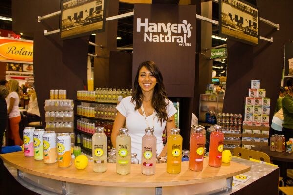 A smiling woman stands behind a curved counter lined with bottles and cans of Hubert's Lemonade beneath a Hansen's Natural sign.