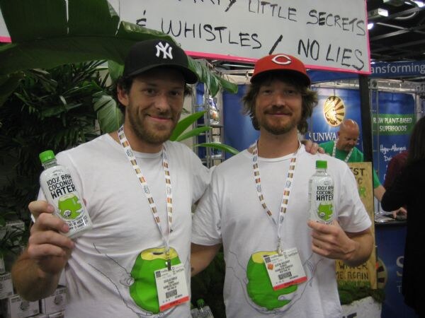 Two men in white t-shirts and baseball caps, one in a Yankees cap and one in a Reds cap, each hold a bottle of 100% raw coconut water in front of green foliage.