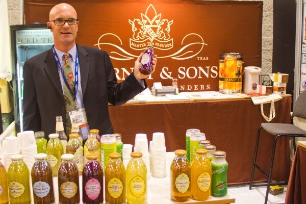 A bald man with glasses in a dark suit holds up a bottle of iced tea at the Harney & Sons booth, with rows of bottled teas on the table in front of him.