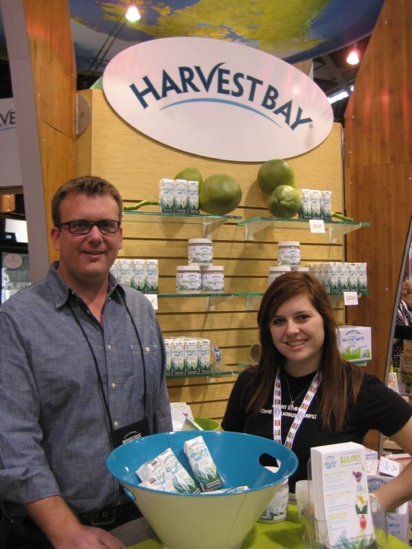 A man with glasses and a woman with a lanyard stand at the Harvest Bay booth, with coconut water cartons and jars on shelves behind them and a bowl of cartons in front.