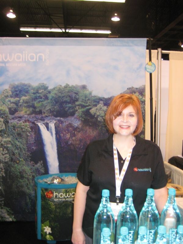 A smiling woman with short red hair in a black polo stands in front of a waterfall backdrop, with bottles of Hawaiian Springs artesian water on the table before her.