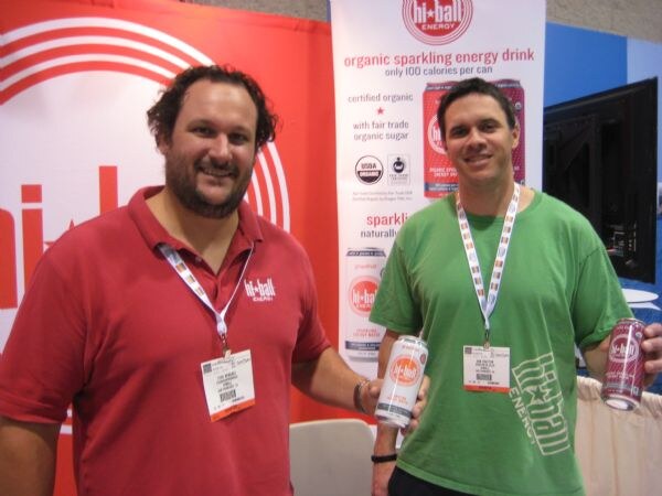 Two men, one in a red Hi Ball Energy shirt and one in a green shirt, each hold a can of sparkling energy drink in front of a Hi Ball Energy banner.