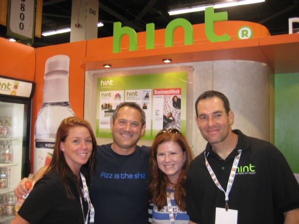 Four people pose together under a green Hint sign; one man wears a shirt reading "Fizz is the shiz," and a display behind them shows a BusinessWeek page.