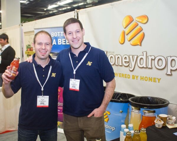 Two smiling men in navy polo shirts with badges stand in front of a Honeydrop banner. One holds up a bottle of orange drink. Jars of drinks sit on a table at right.