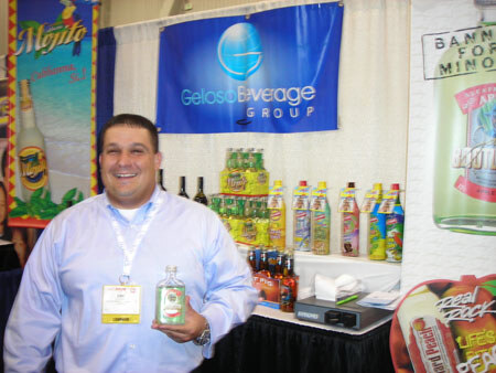 A smiling man in a light blue shirt and badge holds a small bottle in front of a Gelosa Beverage Group banner, a Mejito poster and shelves of bottled drinks.