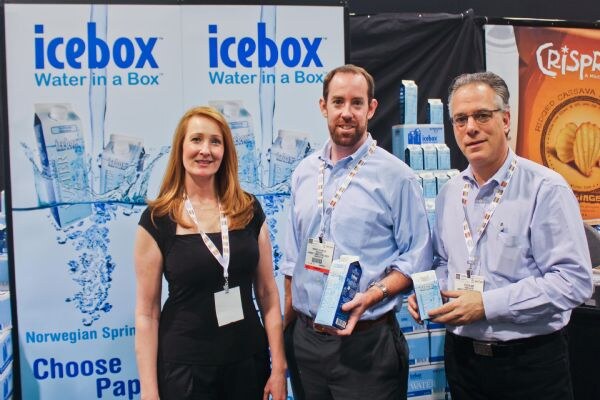 A woman in a black top and two men in light blue shirts stand in front of Icebox Water in a Box banners. The two men hold small cartons. A Crispy Green sign is visible behind them.
