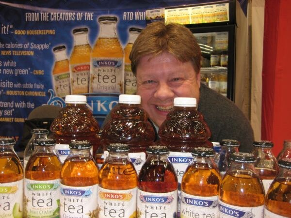A smiling man peers over rows of Inko's White Tea bottles in several flavors, with an Inko's banner behind him and a refrigerated display case at upper right.