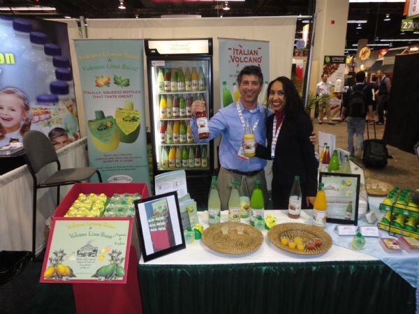 A man in a blue shirt and a woman in a black jacket hold bottles at a booth with Italian Volcano and Volcano Lime Burst banners, a refrigerator of bottled drinks and a table of products.