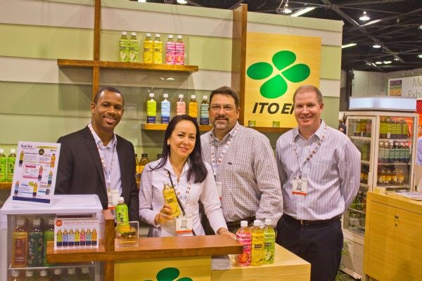 Two men and a woman stand with a third man at a wooden counter in the Ito En booth, with a green clover logo sign and shelves of bottled teas behind them.