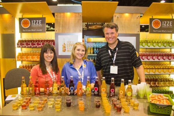 Two women and a man stand behind a counter lined with Izze sparkling juice bottles and plastic sample cups, with Izze signs and shelves of bottles behind them.