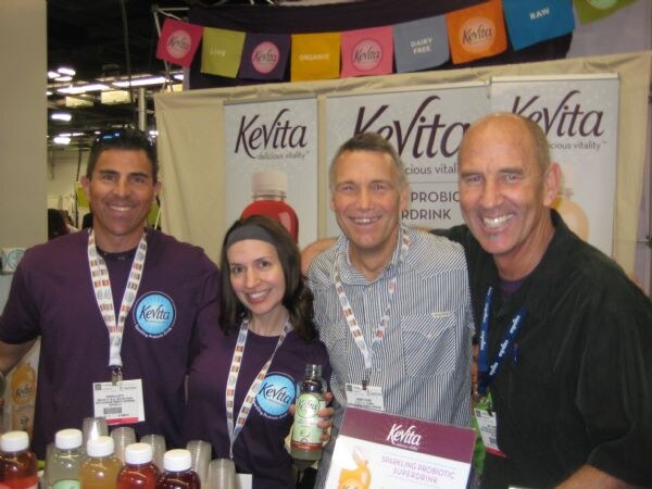 Two men and a woman in purple shirts and a fourth man stand smiling in front of KeVita banners. The woman holds a bottle, and colorful flags hang overhead.