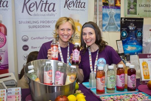 Two smiling women in purple shirts hold KeVita bottles behind a table with bottles in a metal bowl, lemons and a pomegranate, in front of KeVita banners.