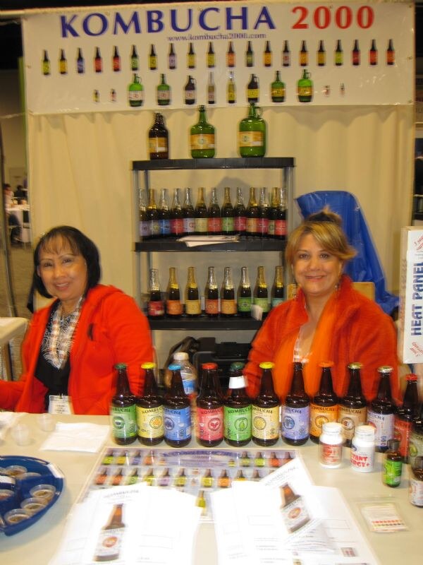 Two women in orange jackets sit behind a table of kombucha bottles with colorful labels. A Kombucha 2000 banner and shelves of bottles are behind them.