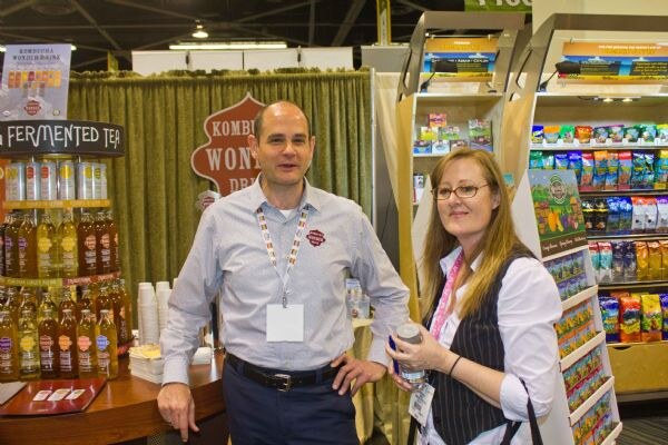 A man in a gray shirt and a woman with glasses holding a bottle stand in the Kombucha Wonder Drink booth, with bottles on a round display and snack shelves nearby.