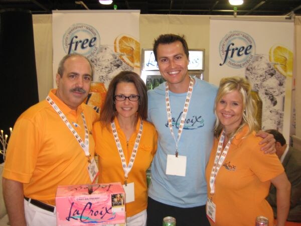 Four people in orange and light blue shirts with lanyards pose in the LaCroix booth in front of banners, with a case of LaCroix sparkling water on the table.