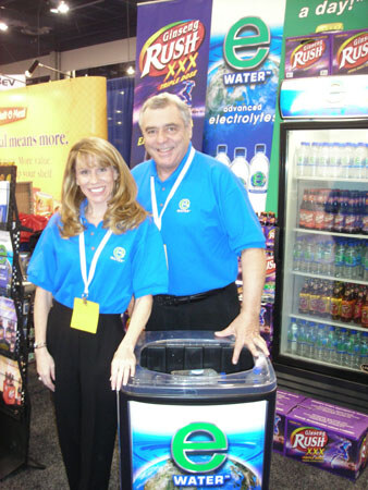 A woman and a man in blue polo shirts stand behind an E Water cooler display, with Glaceau Rush and E Water banners and a refrigerated cooler of bottles behind them.