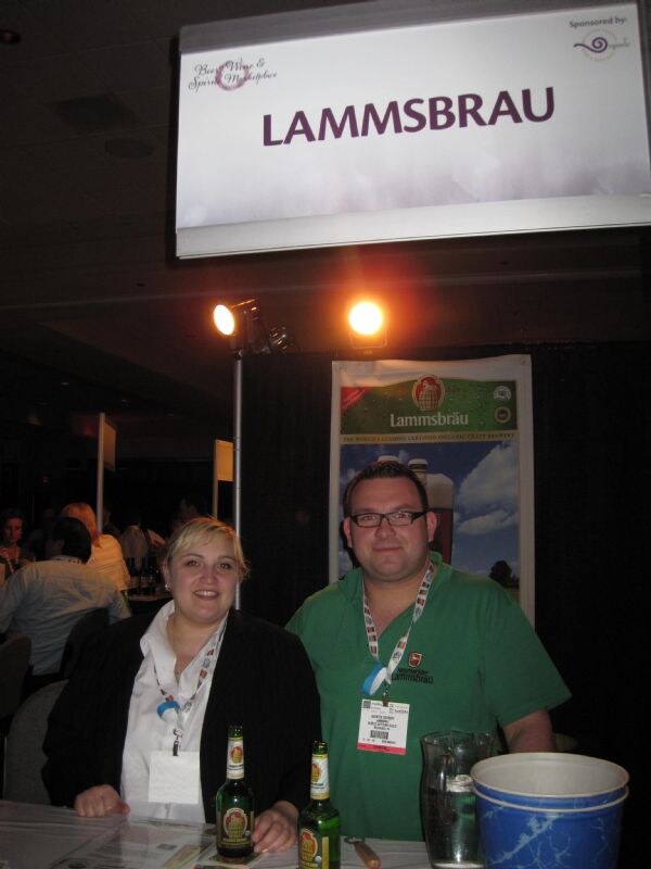 A woman and a man in a green shirt stand behind a table with two beer bottles under a Lammsbrau sign, with a Lammsbräu banner behind them.