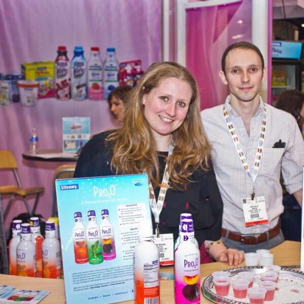 A smiling woman and a man in a checked shirt stand behind a counter with Lifeway Pro2O bottles, a sign and small sample cups of pink drink.