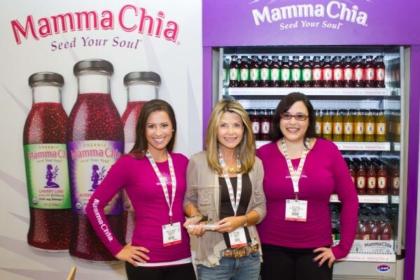 Three women in pink Mamma Chia shirts and a cardigan stand together, one holding a glass award, in front of Mamma Chia bottle graphics and a drink cooler.
