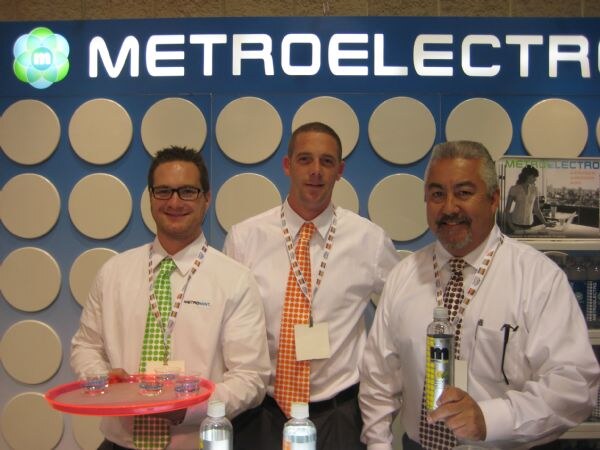 Three men in white shirts and ties stand in front of a Metroelectrolyte sign, one holding a tray of sample cups and another holding a bottle.