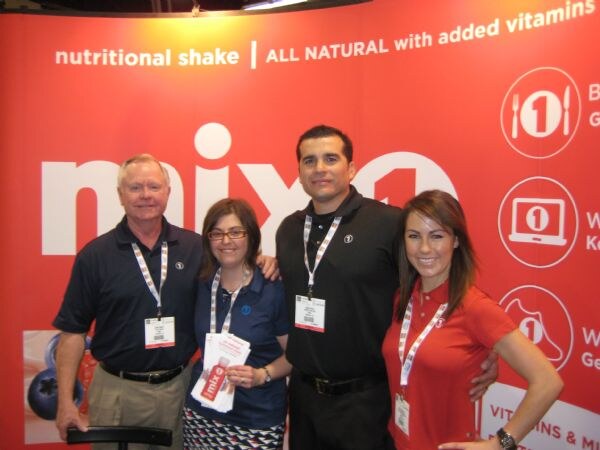 Two men and two women pose in front of a red mix1 banner reading nutritional shake, all natural with added vitamins; one woman holds product flyers.