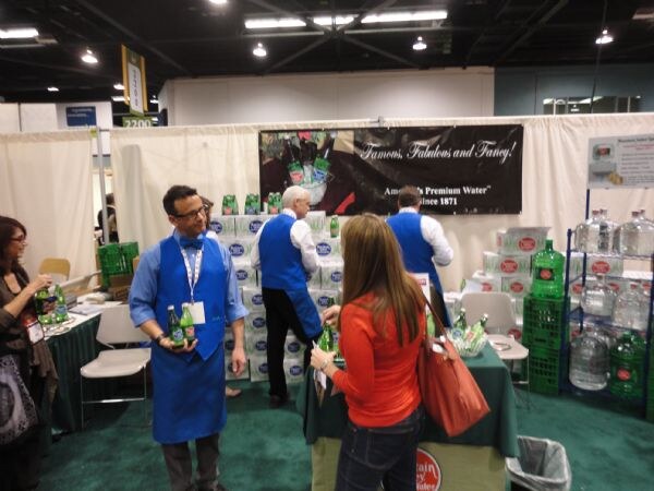 Staff in blue aprons hold green glass bottles in the Mountain Valley Spring Water booth, with stacked cases, large water jugs and a banner reading Famous, Fabulous and Fancy.
