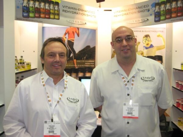 Two men in white MBS shirts with badges stand in the My Body Shots booth, with vitamin-enhanced rehydration shot signs and product shelves behind them.