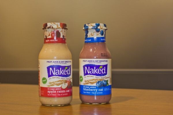 Two Naked bottles on a wooden table: apple raisin oat with a red label and blueberry oat with a blue label, both marked fruit juice and oat smoothie.