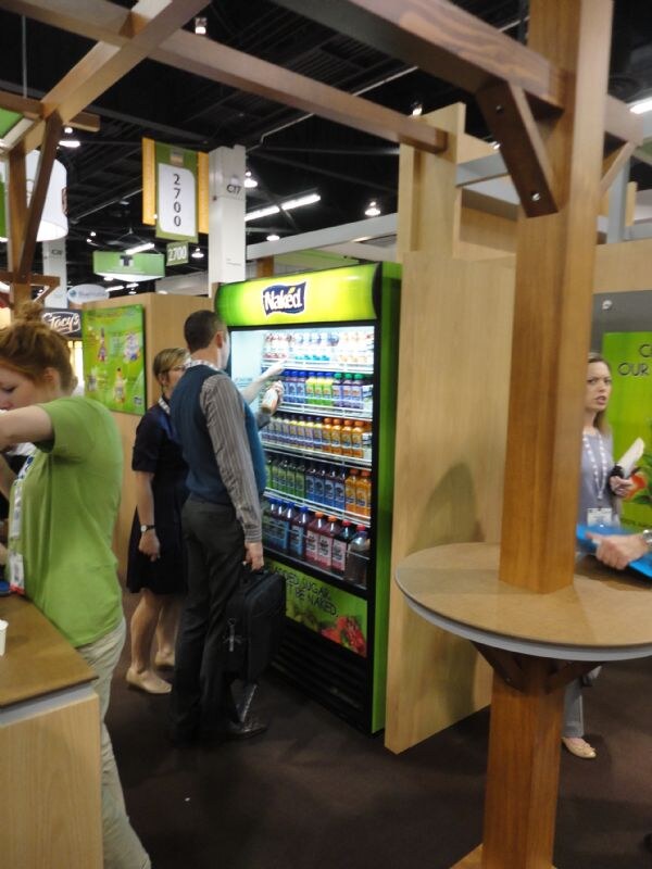 A green refrigerated display case labeled Naked holds bottled juices in a wood-framed booth. A man with a black bag stands at the case, and other attendees are nearby.