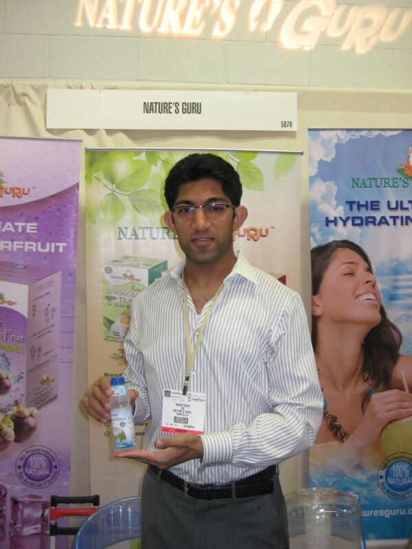 A man in glasses and a striped shirt holds a small bottle of water in front of Nature's Guru banners showing tea and fruit powder boxes. A show badge hangs from his neck.