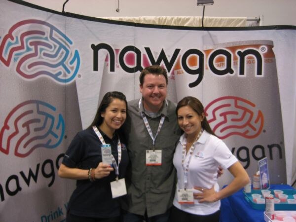 A man stands between two smiling women in front of a nawgan backdrop with a brain logo. All three wear show badges, and drink samples sit on a table at right.