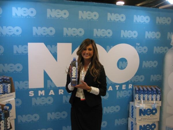 A smiling woman with long hair holds a bottle of NEO Smarter Water in front of a blue NEO backdrop. Cases of NEO bottles sit stacked on both sides.