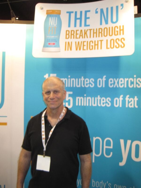 A man in a black polo with a badge lanyard stands beneath a NU FIT sign reading The 'NU' Breakthrough in Weight Loss, in front of a blue display.