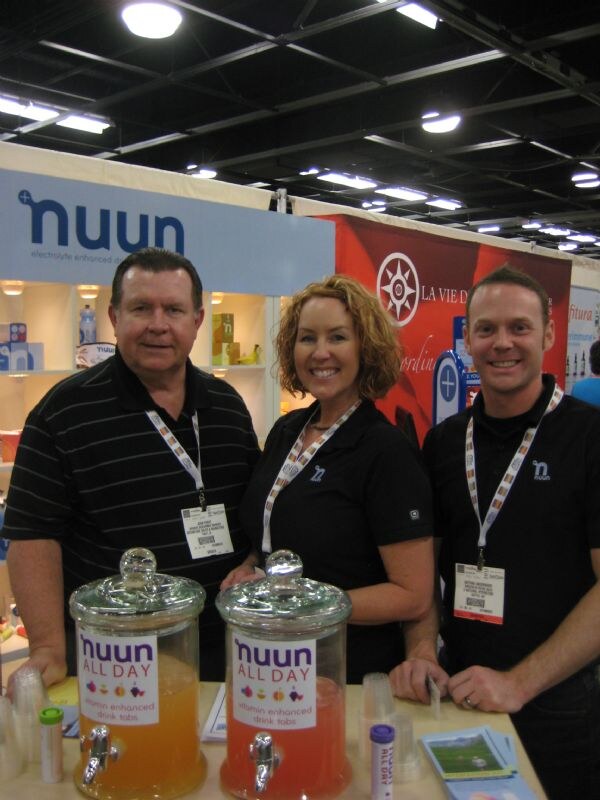 Two men and a woman in black shirts with badges stand behind a counter with two jars of nuun All Day drink. The nuun sign is at left and a La Vie D banner is behind them.